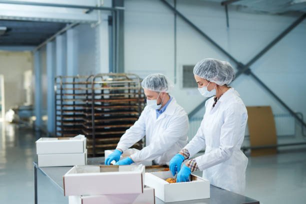 Confectionery factory workers in white coats putting ready pastry into paper boxes.