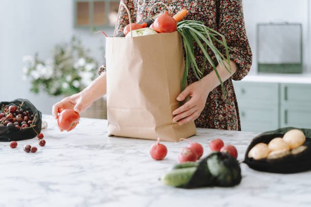 Woman in dress unpacking vegetables from paper grocery bag onto kitchen island with marble top, getting ready to prepare healthy and nutritious meal, furniture in blurred background, cropped shot